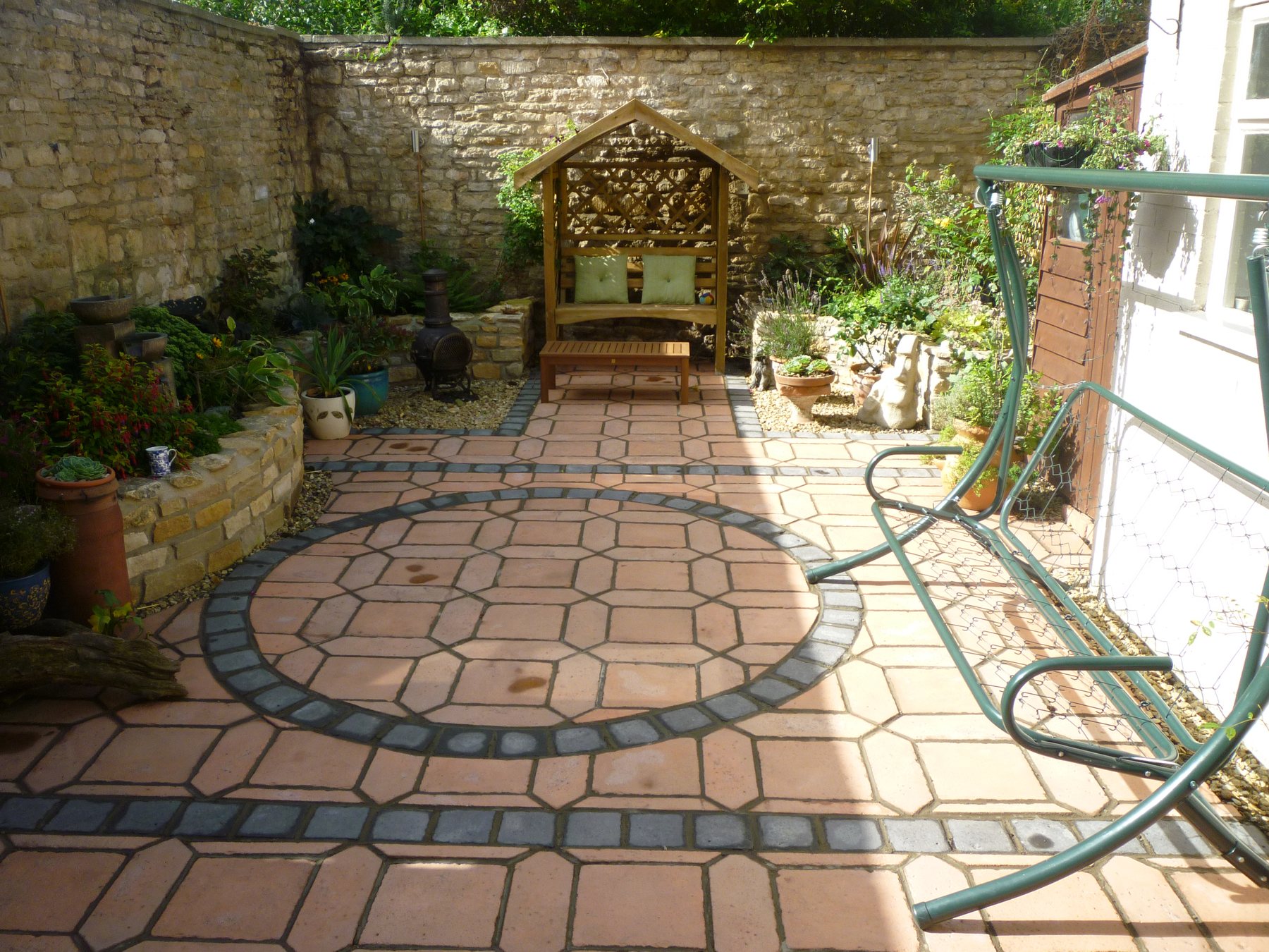 After: A dark corner by a stone wall, now a circle-pattern patio with an arbour seat.